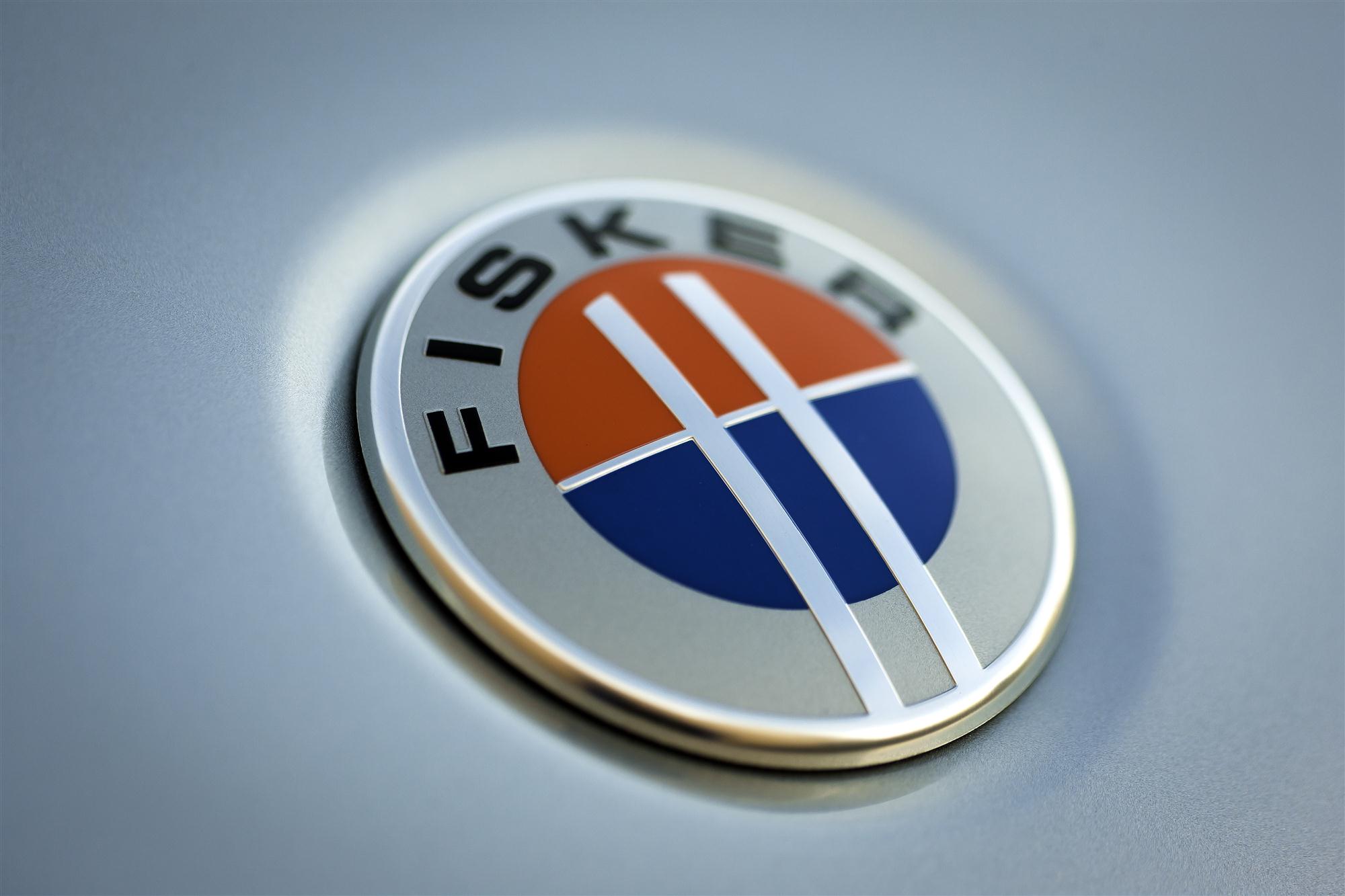 Close-up photo of a Fisker car badge on a vehicle. The badge is silver with a black outline and features the Fisker logo in the center. The logo depicts a stylized trident shape with three prongs.