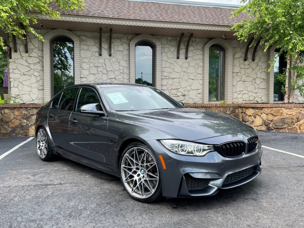 Used 2017 BMW M3 in Gray with sporty alloy wheels, aggressive front fascia, and a sleek design, parked in front of a stone-finished building.