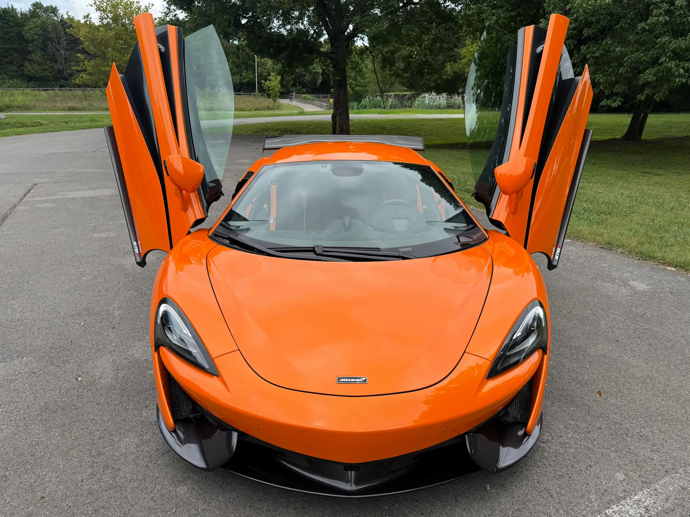 Front view of an orange 2019 McLaren 570S with both dihedral doors raised and its dark front splitter visible.