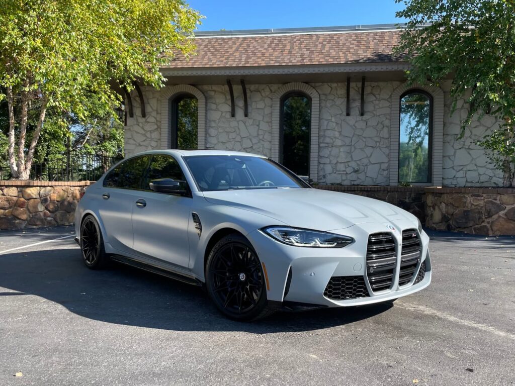 2023 BMW M3 Competition in metallic gray with aggressive front-end styling, sporty alloy wheels, and track-ready performance, parked in front of a stone-finished building.