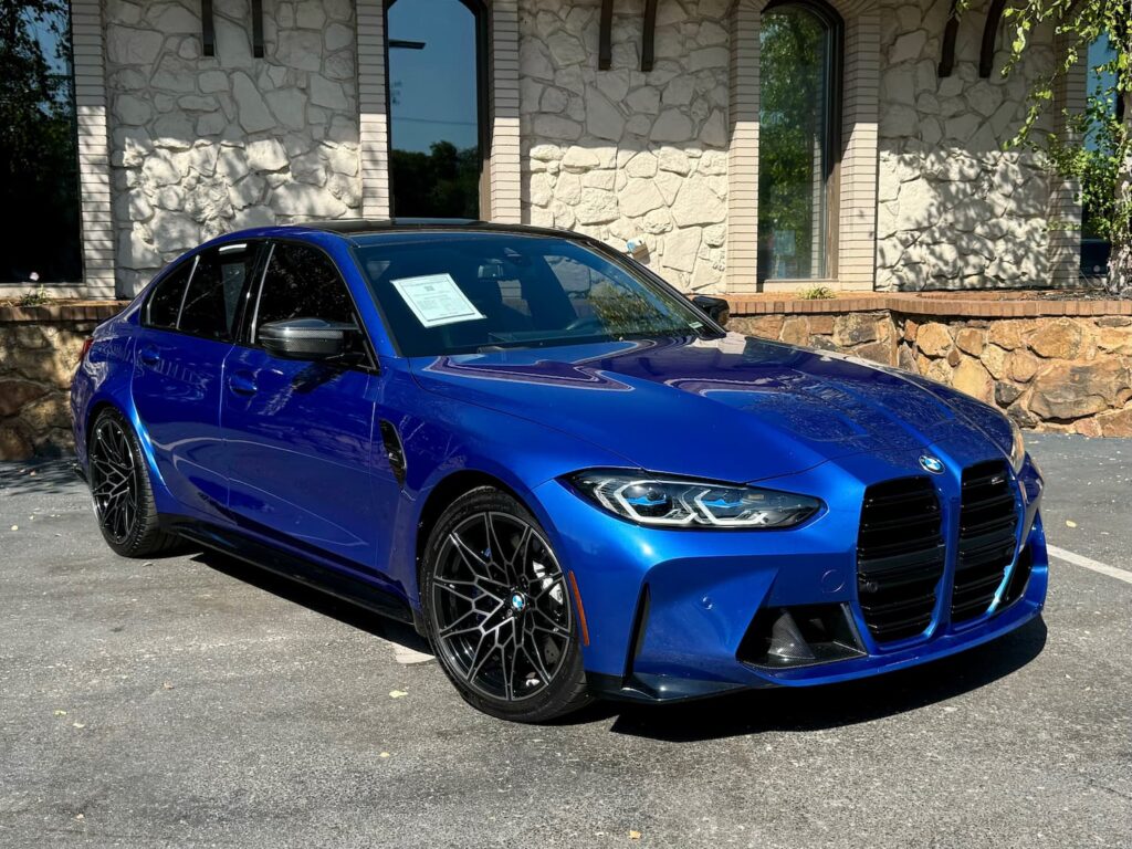2023 BMW M3 Competition in a striking blue finish with black wheels, aggressive front styling, and an aerodynamic body, parked in front of a stone-finished building.