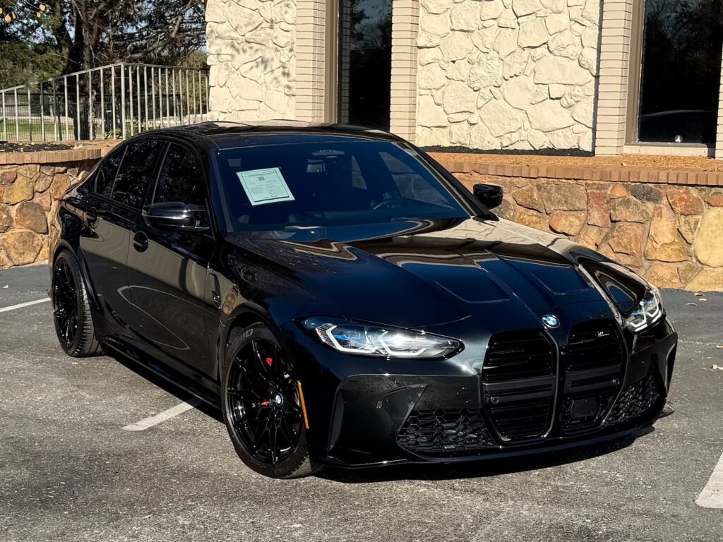 Used 2023 BMW M3 in sleek black with sporty alloy wheels, aggressive front grille, and high-performance design, parked in front of a stone-finished building.