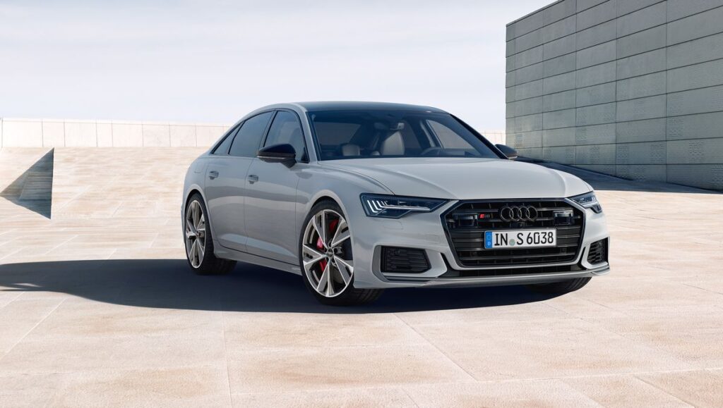 A white Audi S6 sedan parked on a concrete surface in a city. The sun is shining and the sky is blue.