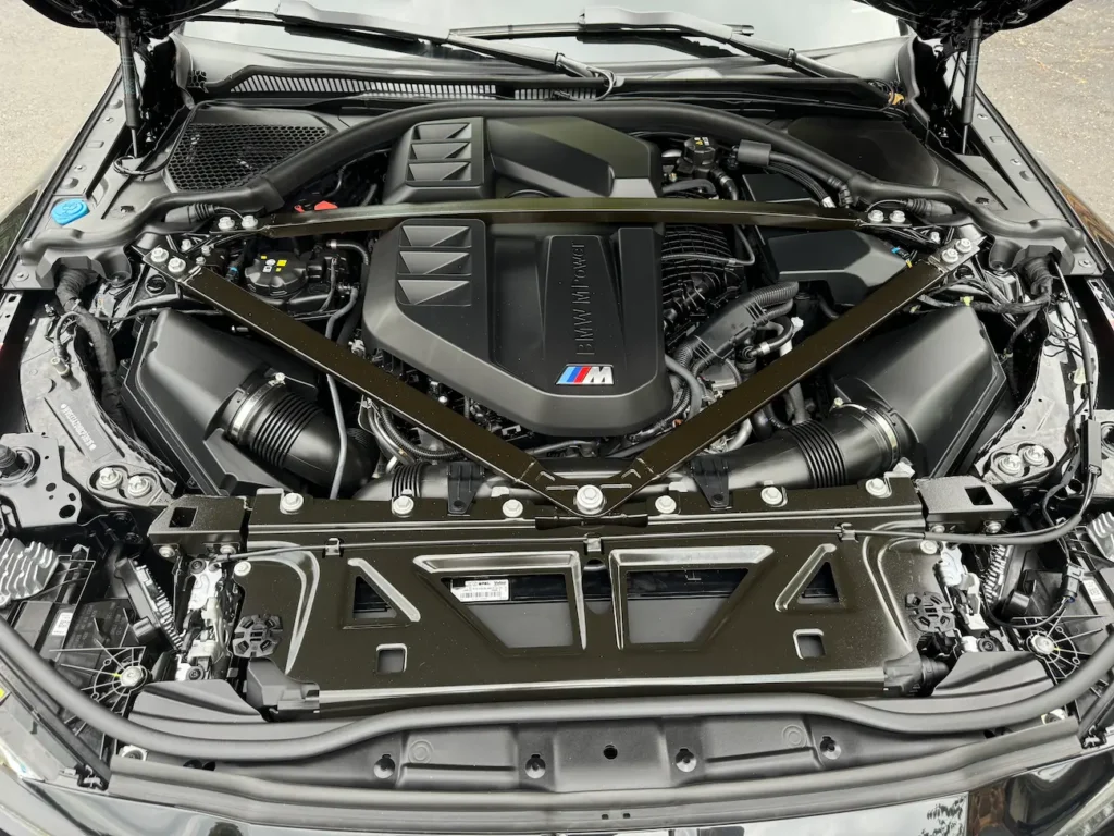 Engine bay of 2024 BMW M4 Competition showing M TwinPower Turbo inline-six engine with carbon strut brace.
