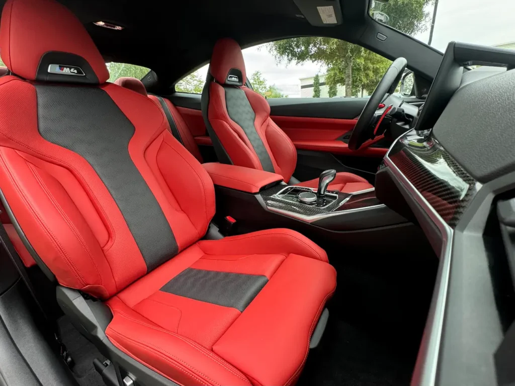 Interior of 2024 BMW M4 Competition with red and black M Sport seats and carbon fiber trim.
