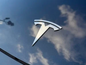 Tesla emblem close-up on gray hood of 2024 Model 3 with sky reflection.