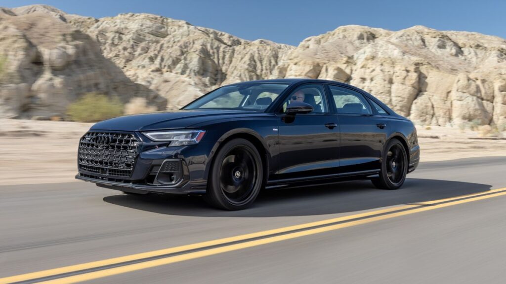 A black Audi A8 sedan driving on a desert road with snow-capped mountains in the background.