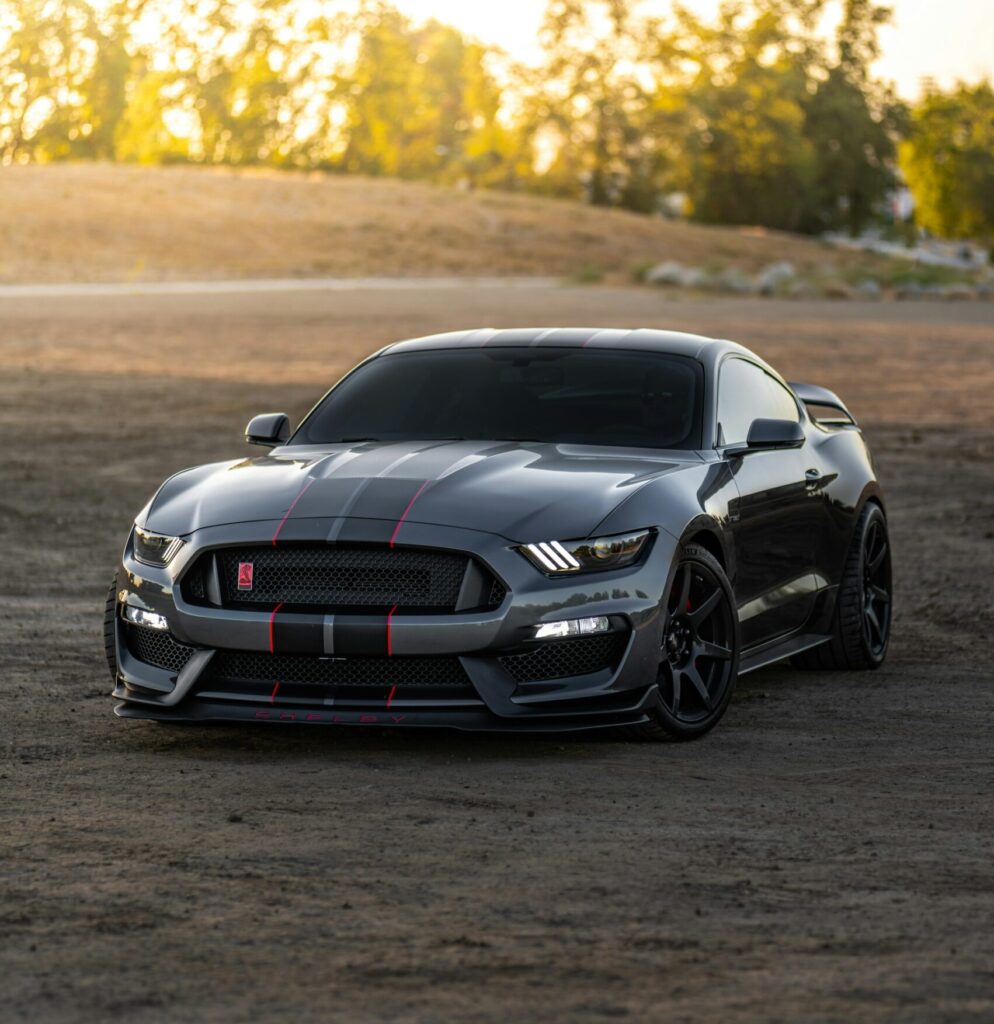 A gray Ford Mustang Shelby GT500 parked on a dirt road with a field and trees in the background.