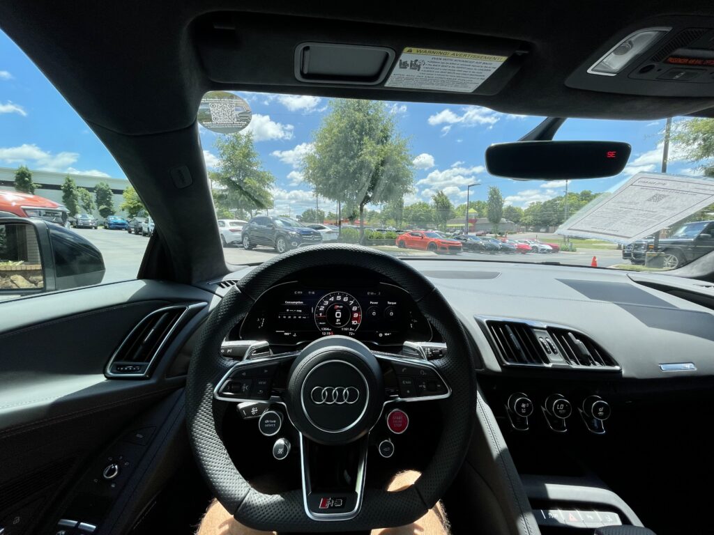 Driver's perspective of a used Audi R8 V10 Plus interior, showcasing the steering wheel, digital dashboard, and a glimpse of the dealership outside.