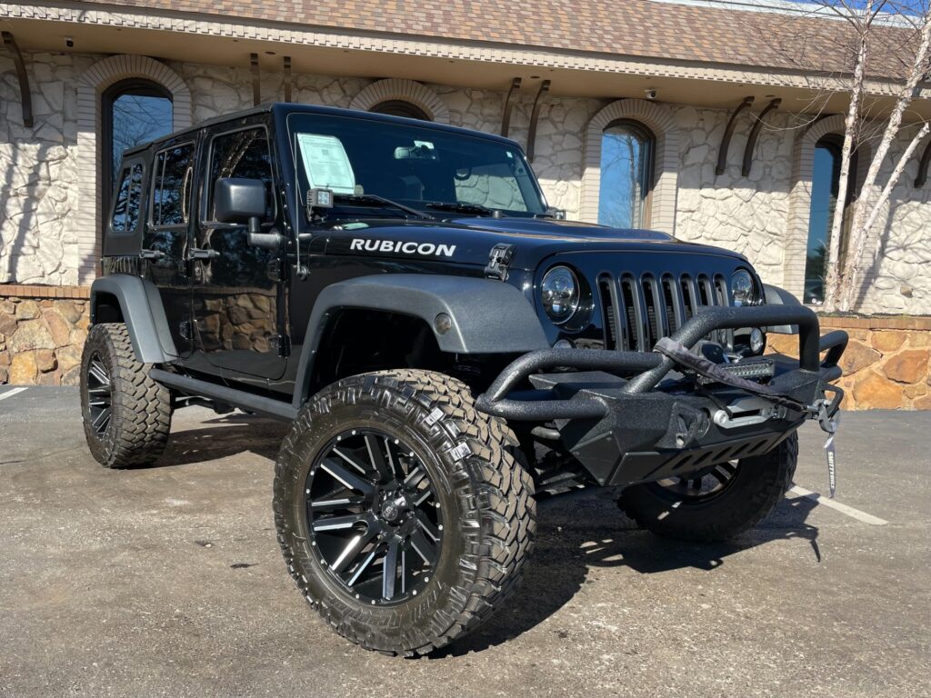 A black Jeep Wrangler Rubicon with a black soft top is parked in front of a stone building with a large glass window and a black metal awning.