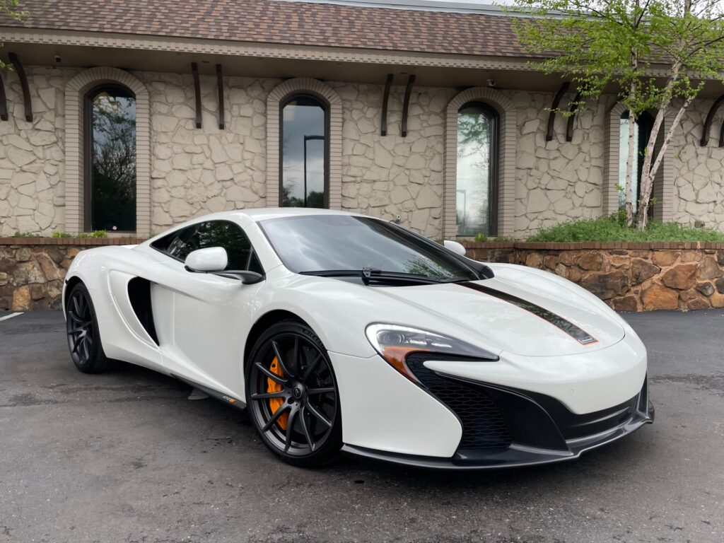 A sleek white 2015 McLaren 650S Coupe with a black interior sits on the showroom floor at AutoPro Nashville, a used car dealership. Sunlight streams through the showroom window and highlights the car's curves.