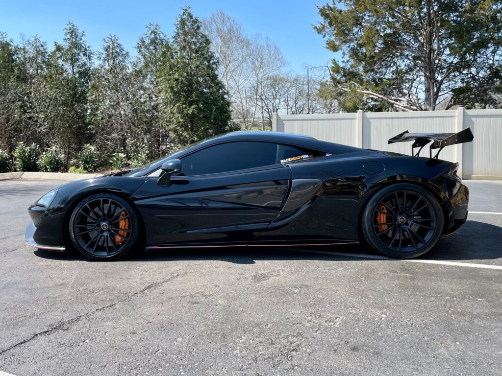 A black McLaren 570S with aftermarket modifications, including a rear wing and side skirts, parked in a parking lot.