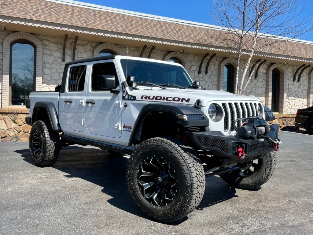 A white Jeep Gladiator Rubicon with aftermarket wheels and a lifted suspension is parked in front of a brick building with a metal awning. The Jeep has a black grill and bumper. There is a "Jeep" and "Rubicon" logo on the vehicle.