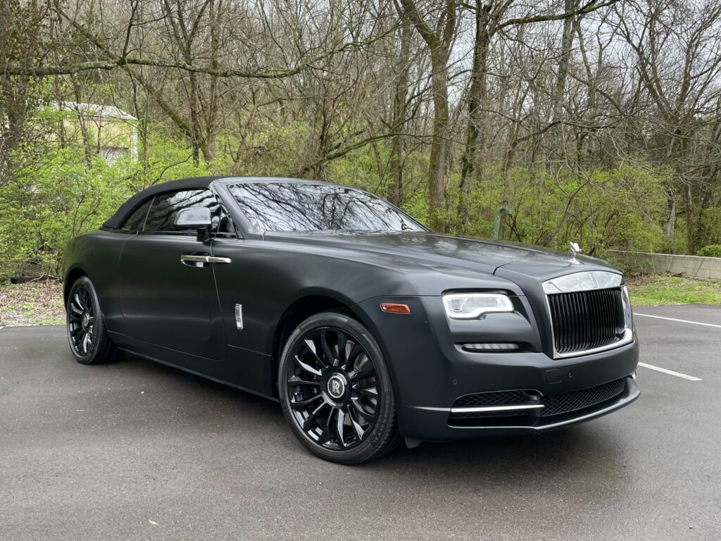 A black matte Rolls-Royce Dawn parked in a dealership showroom next to a forest.