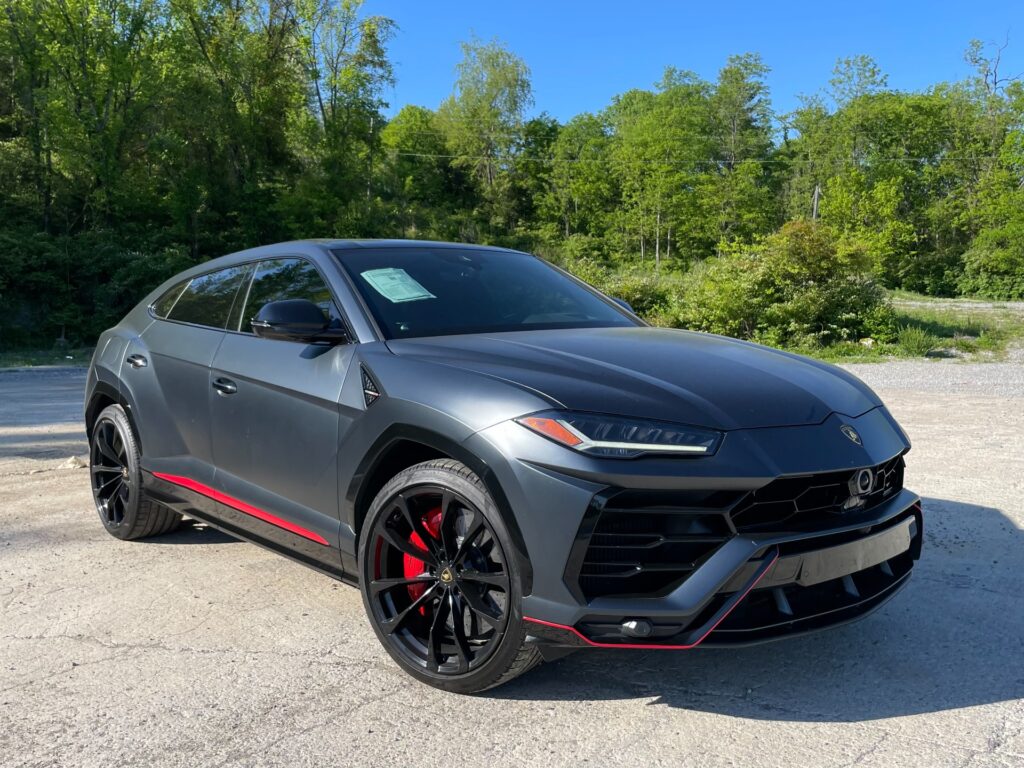 A black Lamborghini Urus, a high-performance SUV, is parked in front of a line of trees at AutoPro Nashville, a used car dealership.
