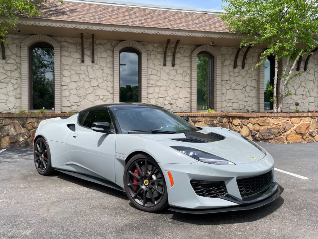 A stylish gray 2021 Lotus Evora GT parked in front of a stone building with a black awning. The high-performance car features a mid-mounted engine and chrome wheels.