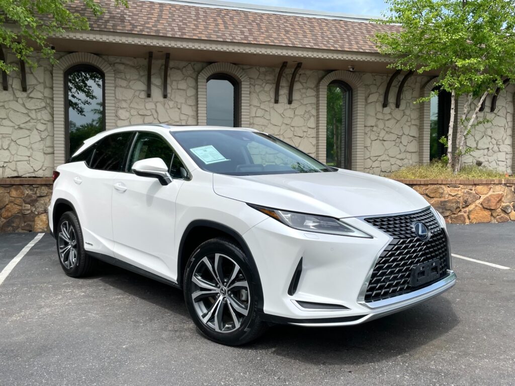 A white 2022 Lexus RX 450h hybrid SUV parked in front of a stone building with a black awning. The Lexus has a sunroof, alloy wheels, and tinted windows.