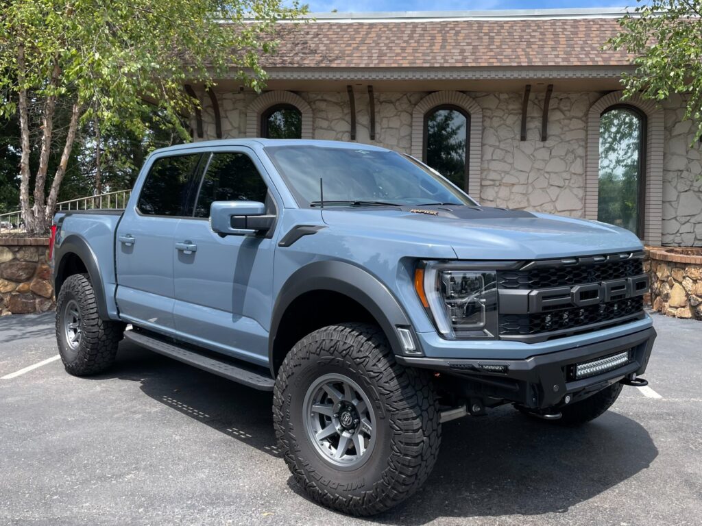 A blue 2023 Ford F-150 Raptor parked in front of AutoPro Nashville's building, with off-road tires and a lifted suspension.