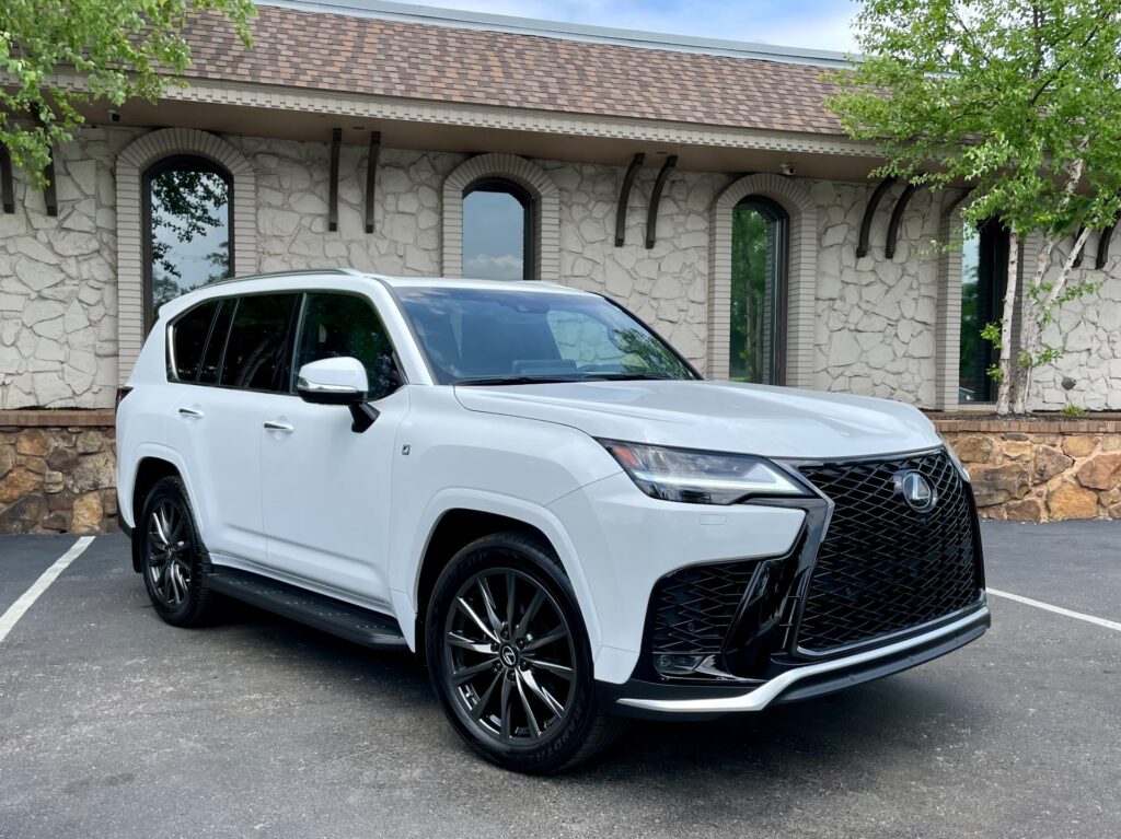 A white 2024 Lexus LX 600 F SPORT luxury SUV parked in front of a stone building with a black awning. The Lexus has a sunroof, chrome grille, and alloy wheels.