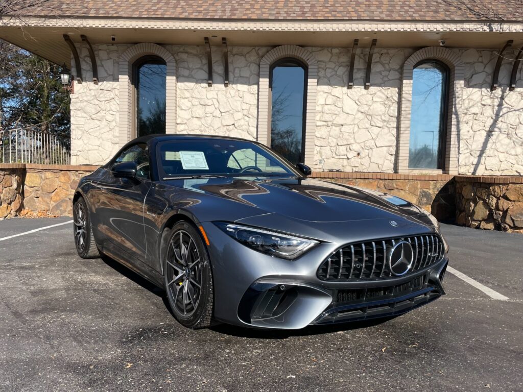 A gray Mercedes-Benz AMG SL 63 convertible parked in front of a stone building with a red awning. The car has the top down and the windows are down. There are trees and bushes in the background.