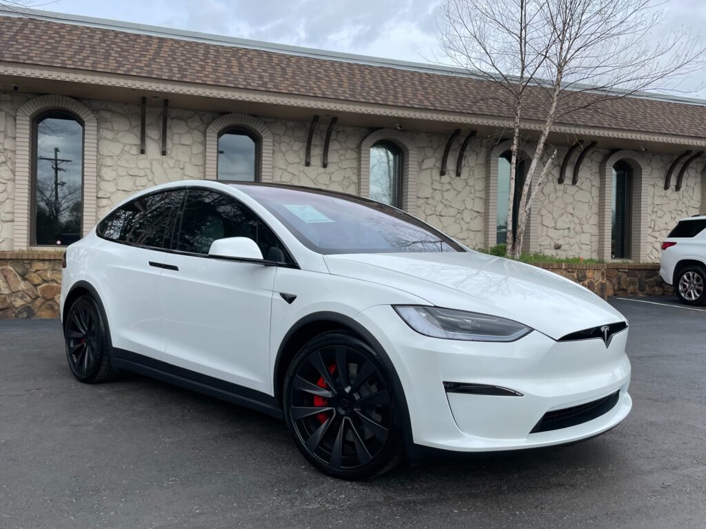 A white Tesla Model X with gull-wing doors open, parked in front of a brick building with a stone roof.