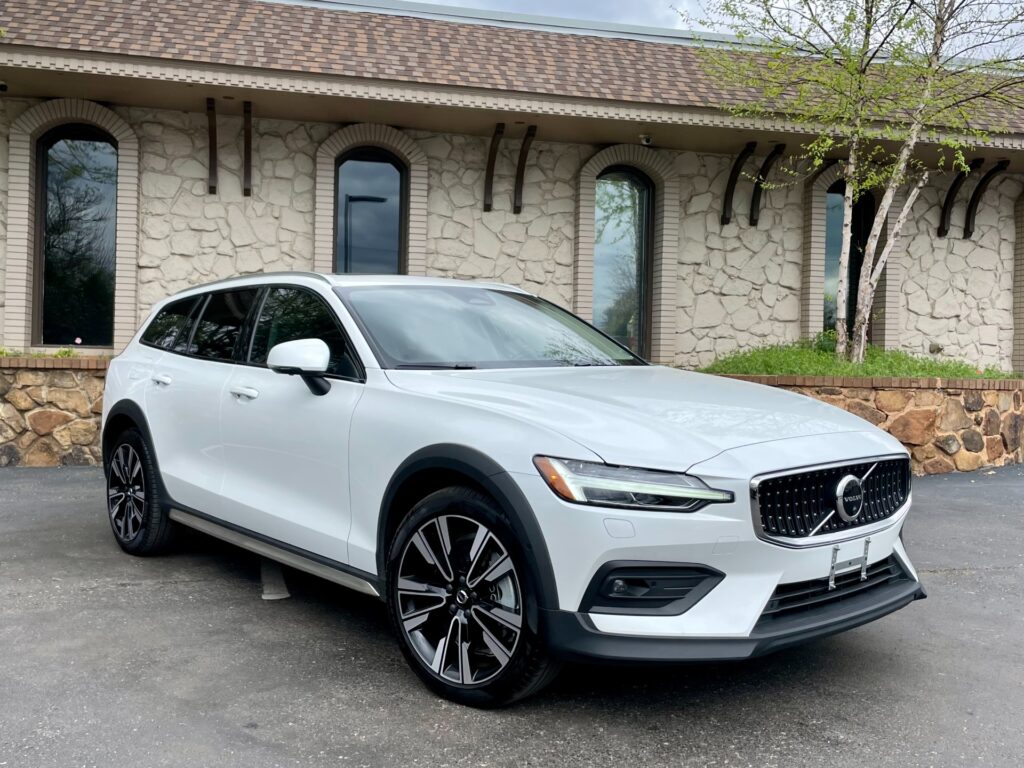 A white Volvo V60 Cross Country wagon is parked in front of a stone building with a large glass window and black awning.
