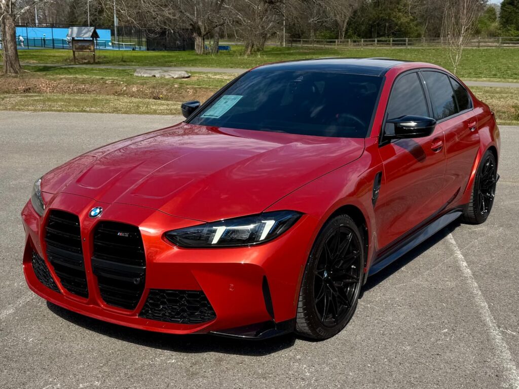 2025 BMW M3 in a striking orange special-order color with black accents, aggressive front grille, and aerodynamic body design, parked outdoors.