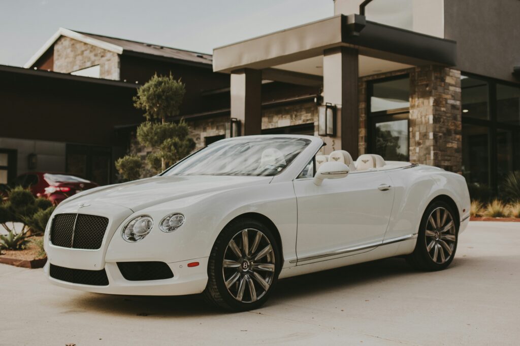 A white Bentley Continental GT convertible parked in front of a modern house with a glass door and windows.