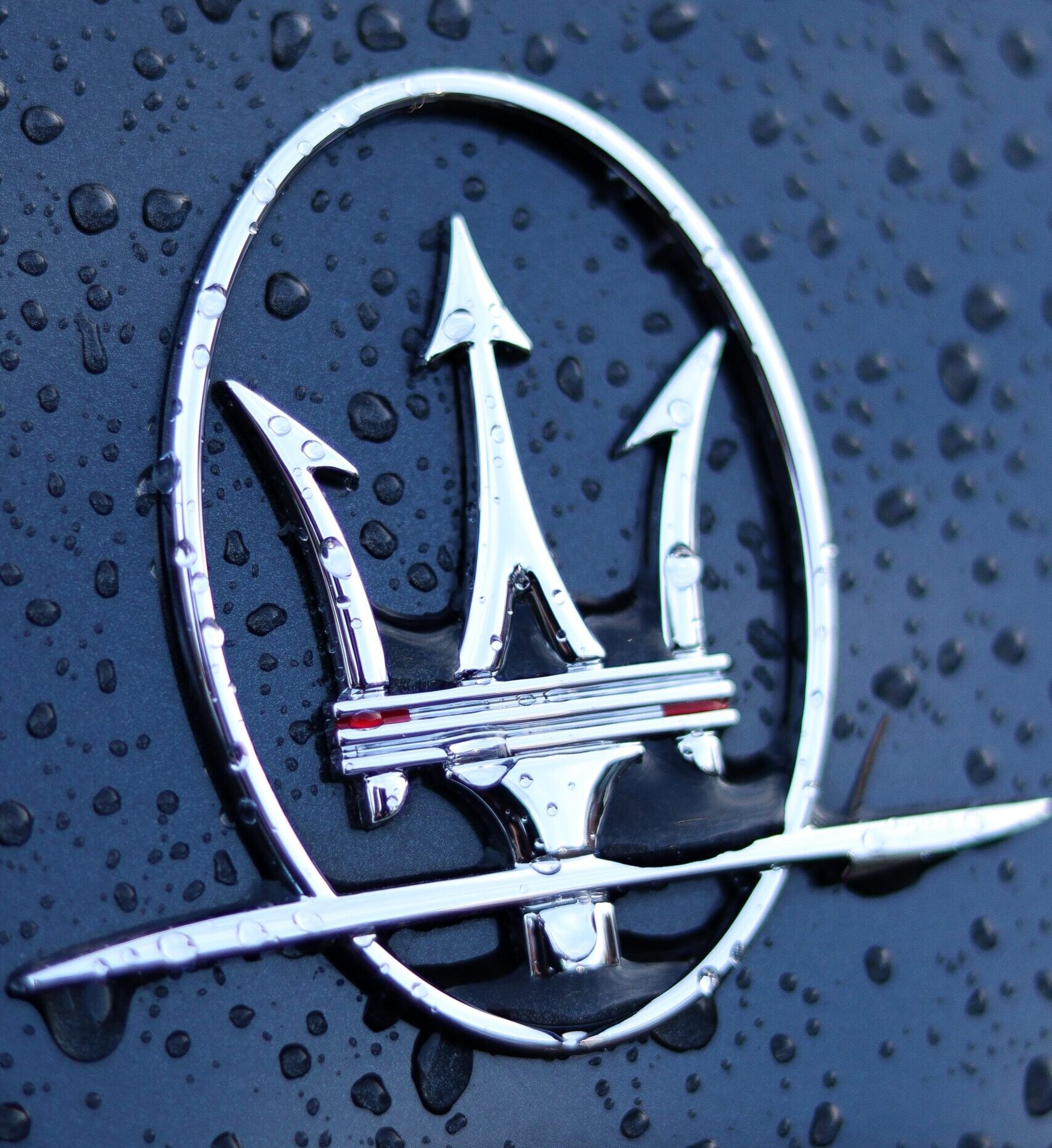 Close-up of the Maserati Trident logo, surrounded by water droplets. The photo highlights the logo's intricate details and the water's reflective qualities.