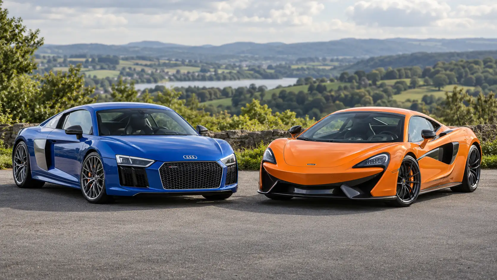 Blue Audi R8 and orange McLaren 570S parked side by side, viewed from the front with green hills behind them.